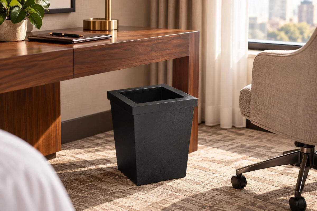 Square black faux leather wastebasket beside a hotel room writing desk
