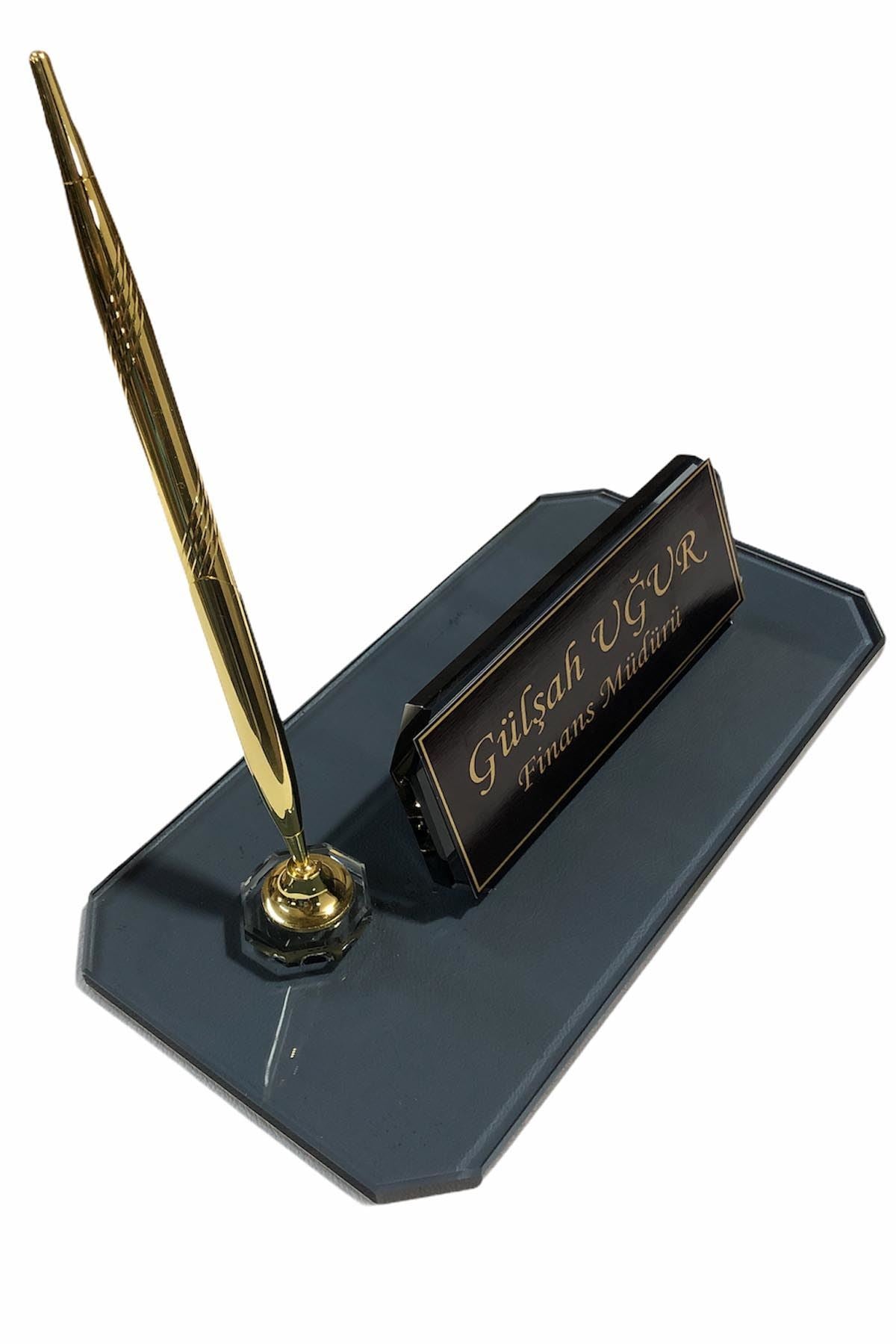 Angled view of a dark glass desk nameplate with a personalized gold-bordered plate and one gold-tone pen